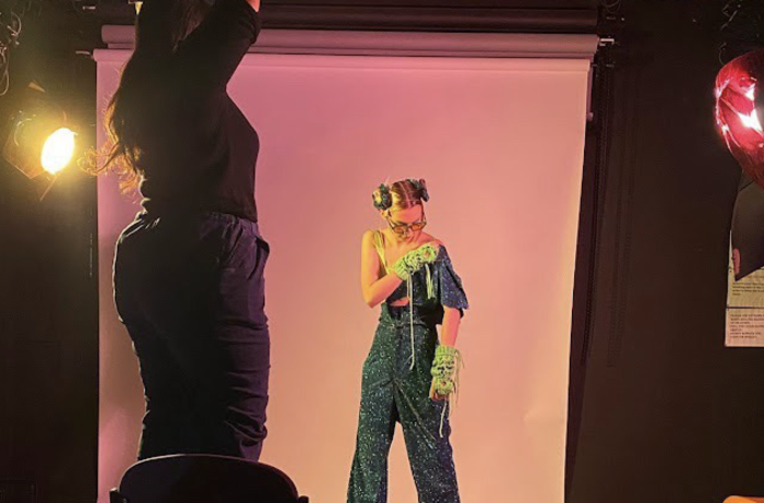 A model stands at the centre of the image wearing a blue outfit with a white backdrop
                    behind her and the lighting is pink. Someone stands to the left wearing all black and they are
                    adjusting the lighting.